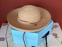 Light tan natural raffia sun hat with wide brim on a purple-flower patterned table and red background, showing full hat shape and leather chin strap.