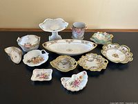 19 assorted vintage porcelain pieces arranged on a table showing various floral and gold decorated dishes and bowls.