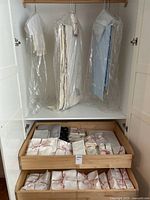 Full wardrobe shot showing hanging tablecloths in dry cleaning bags and two drawers with bundled linens and napkins tied with pink ribbon.
