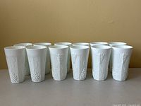 Front view of twelve white milk glass tumblers in a row showing Harvest Grape pressed pattern of grape clusters and leaves.