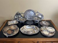 Full set of vintage silverware laid out on black surface showing variety of trays, bowls, platters, and serving baskets with varying tarnish levels.