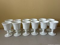Front angle photo of all 12 white milk glass wine glasses arranged in two rows showing the grape and leaf embossed design and size.