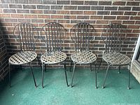 Set of four black wrought iron garden chairs with lattice seats and arched backs shown in a row on green carpet.