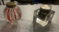Two antique inkwells on a marble surface, one cube-shaped cut glass with silver lid, other tri-footed glass with brass lid and painted stripes