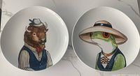 Two porcelain plates on a white surface showing full animal illustrations of buffalo and frog dressed formally