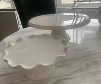 Two white ceramic pedestal dishes on marble table. One larger with embossed dotted rim, one smaller with scalloped edge.