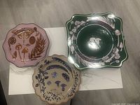 Set of four ceramic plates on white table: three with scalloped edges featuring botanical and mushroom motifs in pink, beige, and blue; one square-edged green plate with white botanical designs.