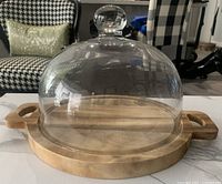 Full view of cheese board with glass dome cover on top, showing wooden handles and round shape.
