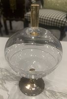Clear spherical glass dish with matching lid on a gold-colored stem and base shown from a slight angle, display on marble surface with checkered background.