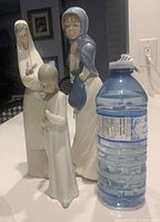 Lot showing three Lladro porcelain figurines of girls in different poses placed next to a water bottle for scale; white, blue and pastel colors, made in Spain.