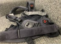 Full view of dark grey BabyBjorn Harmony baby carrier showing breathable mesh fabric, shoulder straps, waist belt, and buckle mechanisms on grey upholstery.