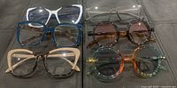 Six pairs of new eyeglasses laid out on black surface showing a range of frame shapes and colors including oversized and round styles.