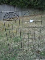 Two black metal garden trellises standing upright on grass against a wooden fence, showing their full height and shape.