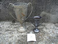Two antique trophies shown side by side on concrete ground. Larger trophy is on the left and smaller trophy on the right.