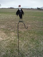 Full view of the metal buzzard mounted on a tall curved metal stake standing in an open grassy field.
