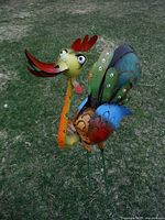 Side angle view showing the multicolored metal rooster with exaggerated eyes, red beak and comb, standing on two green metal legs in a grassy outdoor setting.