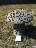 Side view of perforated metal mushroom-shaped light fixture sitting on grass. Shows detailed cutouts, rust, and shape.