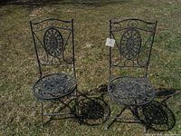 Two black folding wrought iron chairs with intricate circular and oval cutout designs on seats and backrests set on grass. Both chairs show detail of their decorative ironwork and folding function. One chair has a visible repair weld noted on frame.