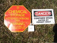 Two safety signs placed on grass, one large red and yellow Chemical Storage octagonal sign and one rectangular Danger Confined Space sign.