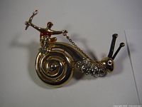 Front view of gold-tone snail brooch with rider holding chain, showing rhinestones and spiral shell detail.
