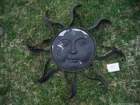 Metal sun decoration laid on grass, showing front with human face and wavy rays, paint wear visible.