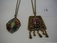 Two necklaces shown together on white background, one silver-tone with multicolored stones, one gold-tone with pink stone and dangles.