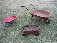 Photo of three items outside on patchy lawn: large rusted wheelbarrow, small red wheelbarrow, rusty planter cart filled with soil and rocks