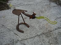 Side angled view of the metal garden sculpture showing shovel blade body, drill bit legs, horseshoe feet, and yellow metal beak.