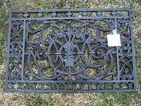 Full top-down view of the rectangular cast iron decorative panel placed on grass, showing intricate scrolling patterns and central floral element.