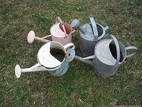 Four vintage metal watering cans shown on grass; includes two plain galvanized cans, one painted pink, and one white with blue leaf motif.