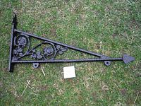 Black metal triangular wall-mounted sign frame with floral designs shown lying on grass, showing full length and detail.