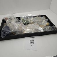 Tray containing various plastic bags filled with assorted costume jewelry pieces including rings, necklaces, bracelets, brooches, and clips.