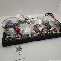 Wide view of assorted costume jewelry pieces in plastic bags arranged in a black tray showing various earrings, bracelets, pins, and small bags containing jewelry.