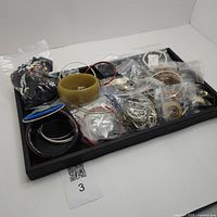 Photo showing a black tray with a variety of costume jewelry including plastic and metal bangles, beaded necklaces, and several plastic bags containing smaller items.