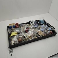 Tray with bags of assorted costume jewelry pieces including necklaces, rings, brooches, pins, cuff links and more, showing a mix of materials and colors.