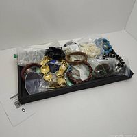 Photo of approximately 2 pounds of assorted costume jewelry in a clear plastic bag placed on a black tray showing various bracelets, necklaces, and smaller bags containing more jewelry