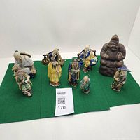 Full shot showing 9 Chinese Mud Men figurines arranged on green felt with large brown Japanese Ebisu statue placed behind them.
