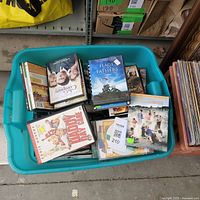 A large green plastic bin filled with assorted DVDs from multiple genres and titles. Visible DVDs include 'Flags of Our Fathers', 'The Day of the Crows', and 'Fearless'.