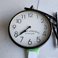 Front view of round clock face with black numbers and Grand Central Station text. Clock mounted on table with mounting bracket visible.