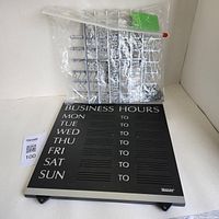 Photo showing the black business hours display board with days of the week printed and several plastic frames with removable silver alphanumeric characters included in a clear plastic bag.
