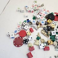 Assorted board game pieces including dice, red tokens, metal miniatures, and green/red plastic buildings scattered on a white surface.