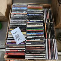 Top-down photo of a cardboard box filled with stacked music CDs in jewel cases with visible titles and labels.