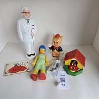 Overview of the lot including Colonel Sanders bank, Marge Simpson figure, Porky Pig figure, Sylvester the Cat in bag, wind-up dog piggy bank, and dog house bank.