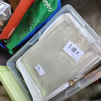 Large translucent plastic storage tote filled with clear plastic sleeves visible on top, some paper bags and plastic bags around.