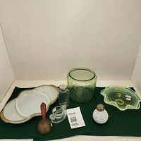 Photo displaying the full lot including a white milk glass divided dish with gold trim, green uranium glass jar, green Fire-King ashtray, brass bell, clear and opaque salt and pepper shakers, and clear glass chicken egg container.