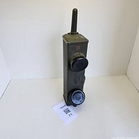 Front view of vintage military walkie talkie showing rectangular metal body, antenna, two black circular speaker/microphone units, and carrying strap.