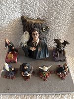 Photo showing all 7 Harry Potter themed fantasy figurines arranged on fabric surface. Central figure of Harry Potter with owl Hedwig on his arm in front of a tree stump. Surrounding figures include Hermione Granger, Hagrid, Dumbledore, and other characters posed on small bases resembling books.