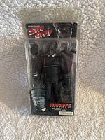 Front view of Sin City Manute action figure in clear blister packaging showing figure and accessories.