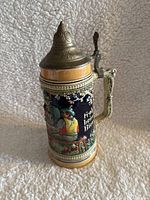 Side view of vintage German ceramic beer stein showing detailed relief artwork of a man with a woman in traditional German dress, pewter lid, and cream/navy color scheme.
