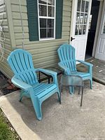 Two blue plastic Adirondack chairs next to two small outdoor tables with glass tops, placed outside near a building with green siding and shutters.